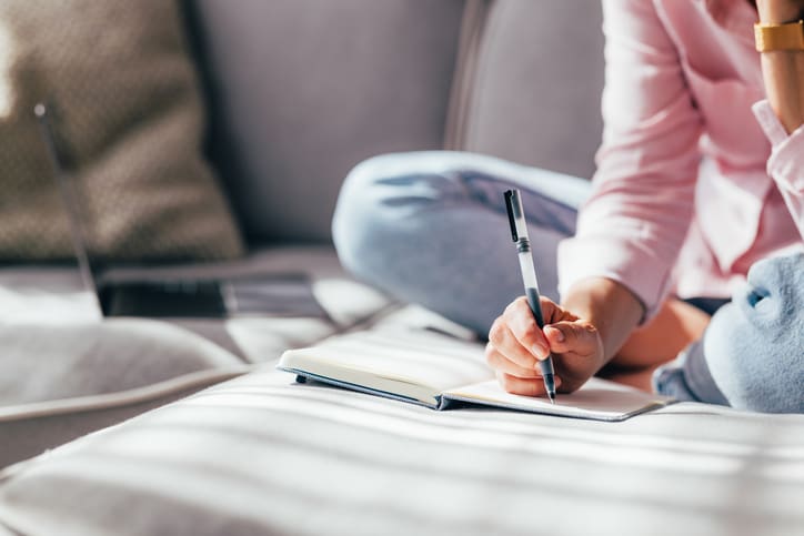 Close-up of a person's hands writing carefully in a blank notebook, sitting comfortably on a couch.