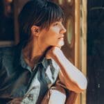 Woman sitting indoors, resting her chin on her hand, looking thoughtfully out a window with soft sunlight illuminating her face.