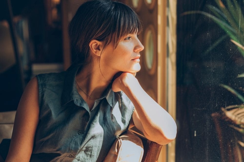 Woman sitting indoors, resting her chin on her hand, looking thoughtfully out a window with soft sunlight illuminating her face.