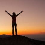 A person stands on a mountaintop at sunrise or sunset with arms raised, symbolizing freedom, empowerment, and personal achievement.