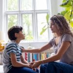 A woman with long curly hair sits on a couch in a bright living room, gently holding a young boy’s hands and looking at him with a caring expression. The boy, wearing a striped shirt, faces her as they talk. Large windows and green plants are visible in the background.