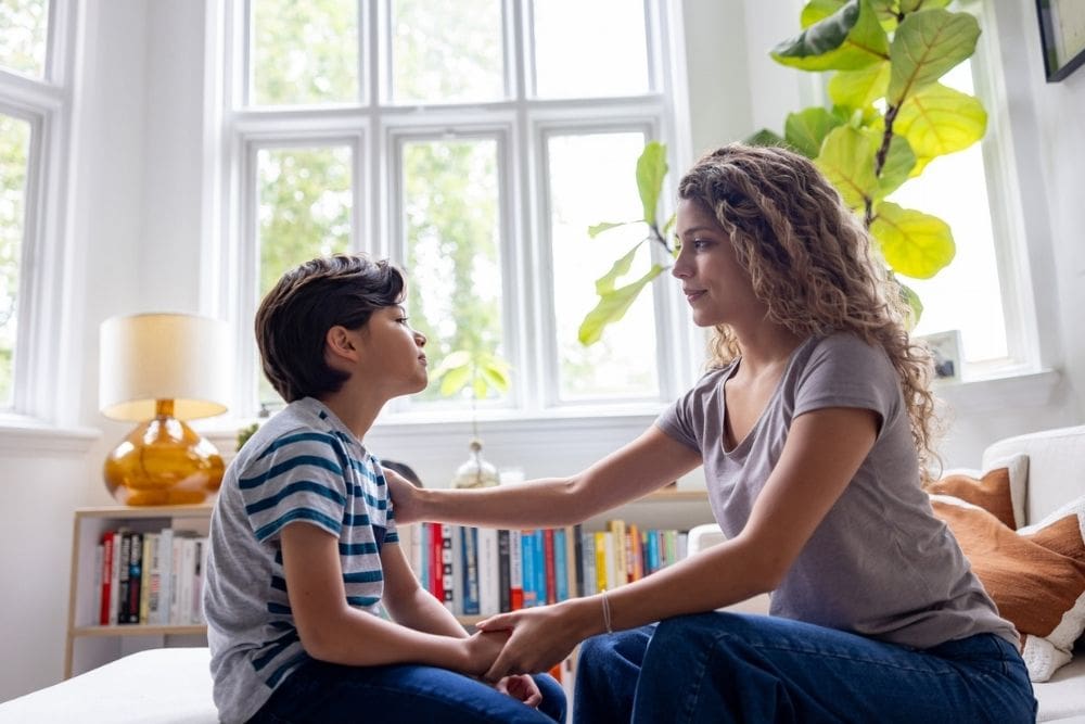 A woman with long curly hair sits on a couch in a bright living room, gently holding a young boy’s hands and looking at him with a caring expression. The boy, wearing a striped shirt, faces her as they talk. Large windows and green plants are visible in the background.
