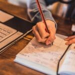Person writing in a paper planner with a pencil next to an open laptop and a smartphone on a wooden desk.