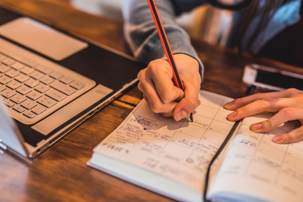 Person writing in a paper planner with a pencil next to an open laptop and a smartphone on a wooden desk.