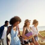 Group of young adults walking together on a sunny beach, smiling and carrying towels and bags, enjoying a carefree summer day by the ocean.