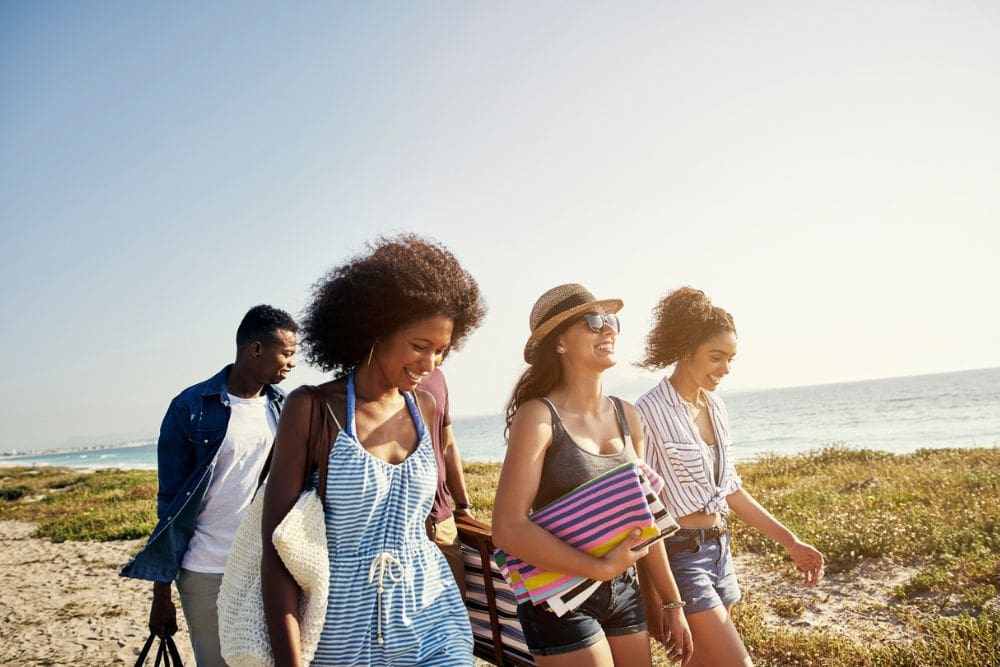 Group of young adults walking together on a sunny beach, smiling and carrying towels and bags, enjoying a carefree summer day by the ocean.