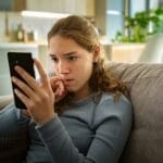 Portrait of a nervous teen girl looking at her smartphone biting her nails worried about bad news