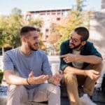 Two men sitting outdoors on a sunny day, smiling and talking openly, symbolizing friendship, support, and meaningful conversation about mental health.