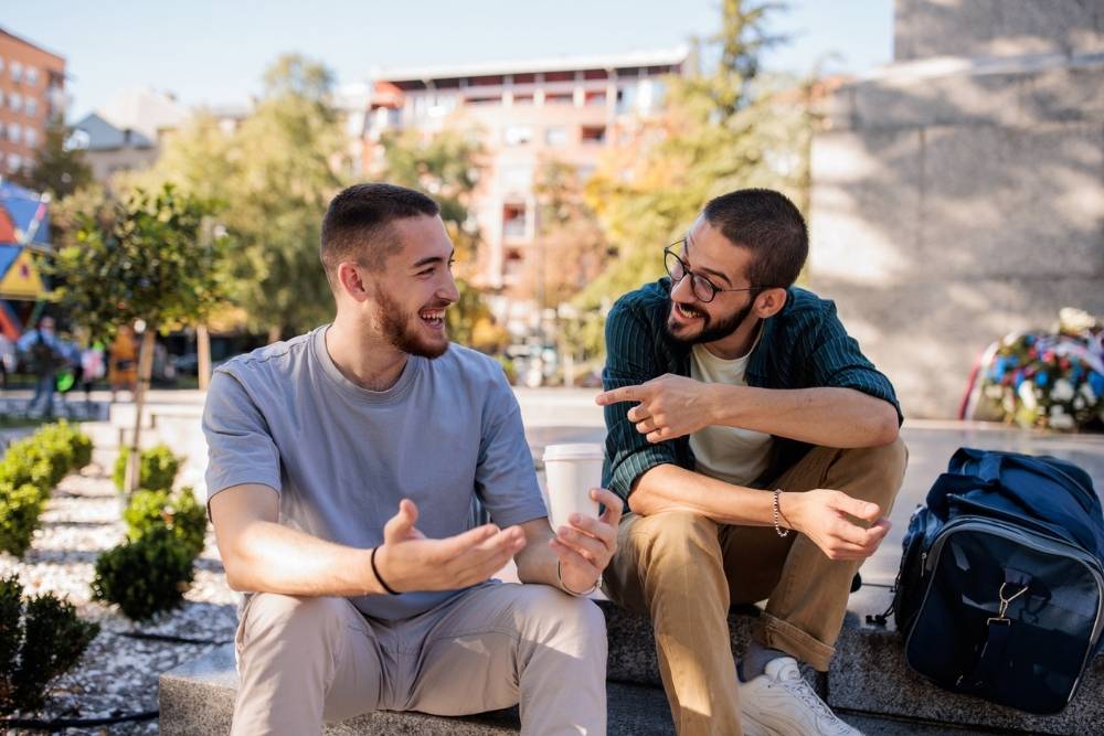 Two men sitting outdoors on a sunny day, smiling and talking openly, symbolizing friendship, support, and meaningful conversation about mental health.