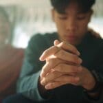 A teenager sits with hands clasped and head down, looking distressed, while an adult rests a comforting hand on his shoulder in the background.