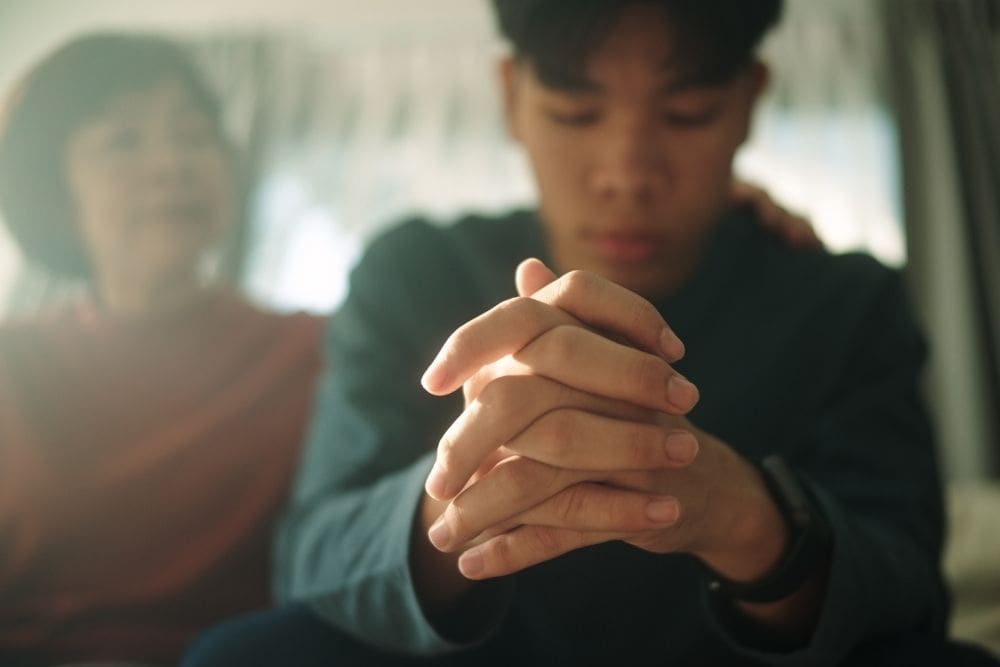 A teenager sits with hands clasped and head down, looking distressed, while an adult rests a comforting hand on his shoulder in the background.