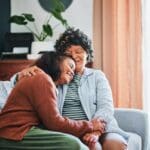 Two women sitting on a couch sharing a warm embrace, smiling and showing love and support in a comforting home setting.