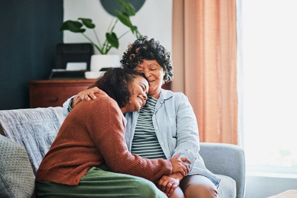 Two women sitting on a couch sharing a warm embrace, smiling and showing love and support in a comforting home setting.