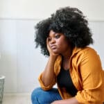 woman looking sad, sitting on the edge of the couch with her head resting on her hand