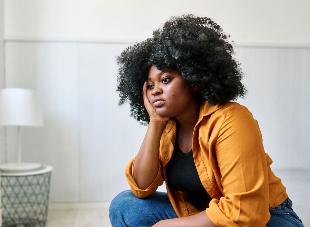 woman looking sad, sitting on the edge of the couch with her head resting on her hand