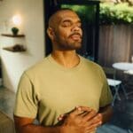 A man stands indoors with his eyes closed, hands resting on his chest, appearing calm and relaxed while practicing deep breathing.