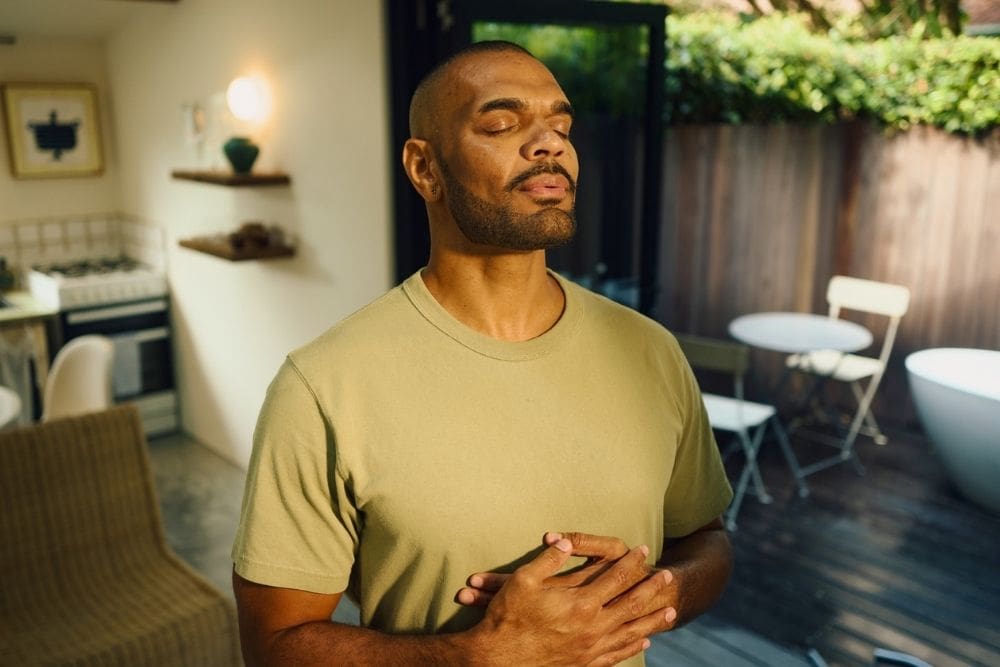 A man stands indoors with his eyes closed, hands resting on his chest, appearing calm and relaxed while practicing deep breathing.