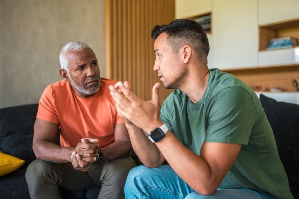 Two men sitting on a couch engaged in a serious conversation, one listening attentively while the other speaks with expressive hand gestures.
