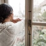 Woman in a cozy white sweater looking out a window, appearing thoughtful and reflective on a cloudy day.