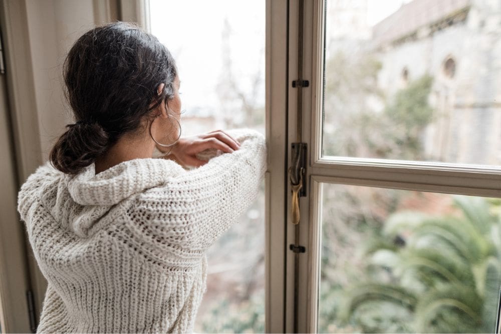 Woman in a cozy white sweater looking out a window, appearing thoughtful and reflective on a cloudy day.