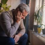 An older man sitting indoors with his head bowed and hand on his face, appearing sad, tired, or overwhelmed.
