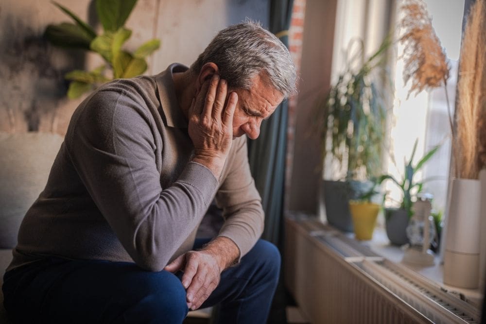 An older man sitting indoors with his head bowed and hand on his face, appearing sad, tired, or overwhelmed.