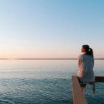 A woman sits on the edge of a pier, gazing out over calm water at sunset, reflecting quietly in a peaceful outdoor setting.