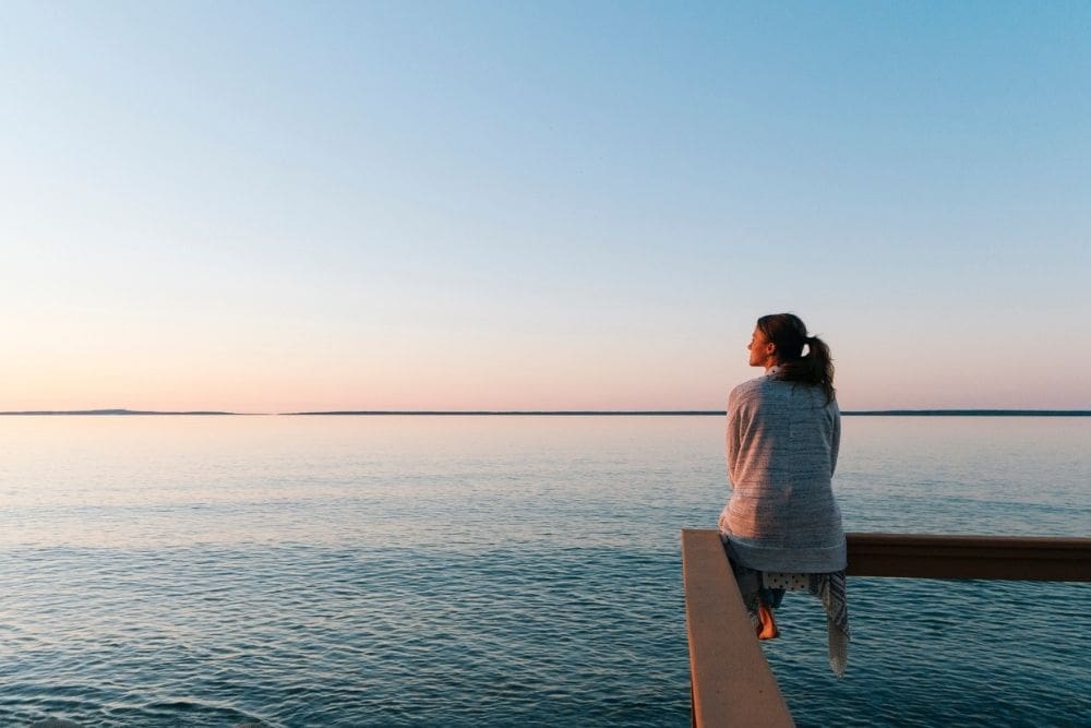 A woman sits on the edge of a pier, gazing out over calm water at sunset, reflecting quietly in a peaceful outdoor setting.