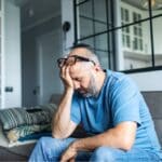 A man sits on a couch with his head resting in his hand, appearing stressed and overwhelmed in a quiet living room.