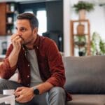 houghtful man sits on a couch holding a coffee mug, looking worried in a bright living room.