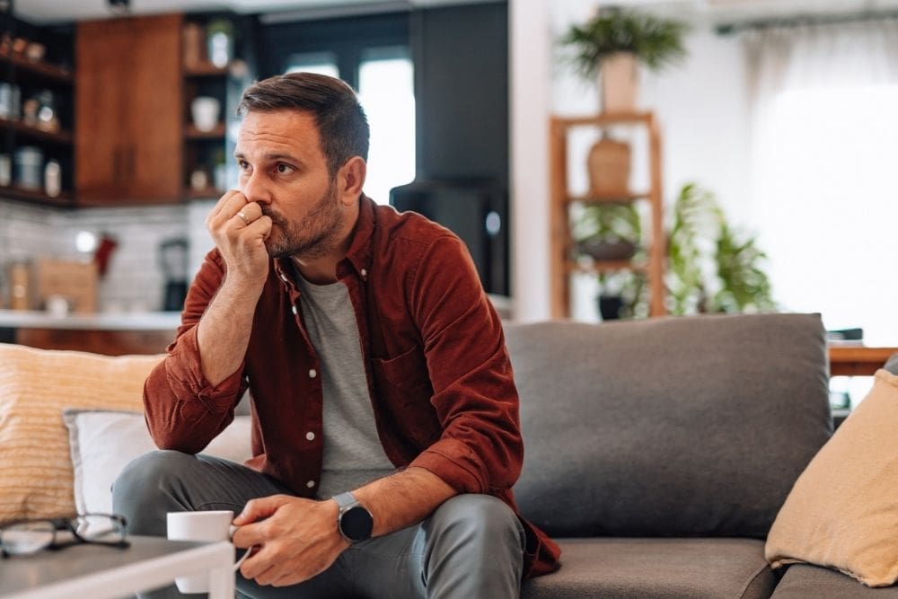 houghtful man sits on a couch holding a coffee mug, looking worried in a bright living room.