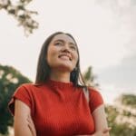 A woman in a red top stands outdoors with her arms crossed, smiling and looking upward, embracing the bright sky and trees—an inspiring reminder to prioritize your mental health.