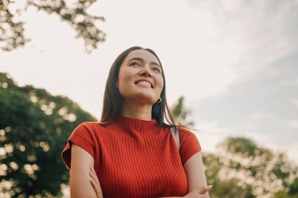 A woman in a red top stands outdoors with her arms crossed, smiling and looking upward, embracing the bright sky and trees—an inspiring reminder to prioritize your mental health.