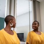 Person smiling at their reflection in a mirror, practicing positive self-talk in a calm indoor setting.